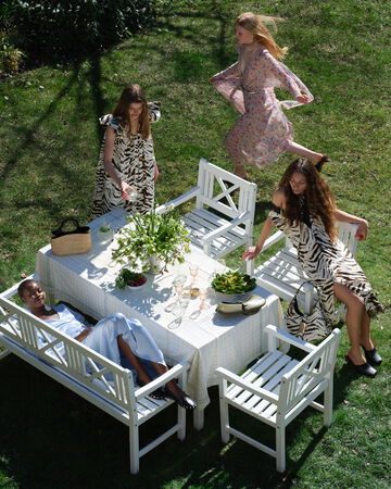 Candid garden party scene featuring guests in various GANNI floral and patterned dresses sitting at a white outdoor dining table
